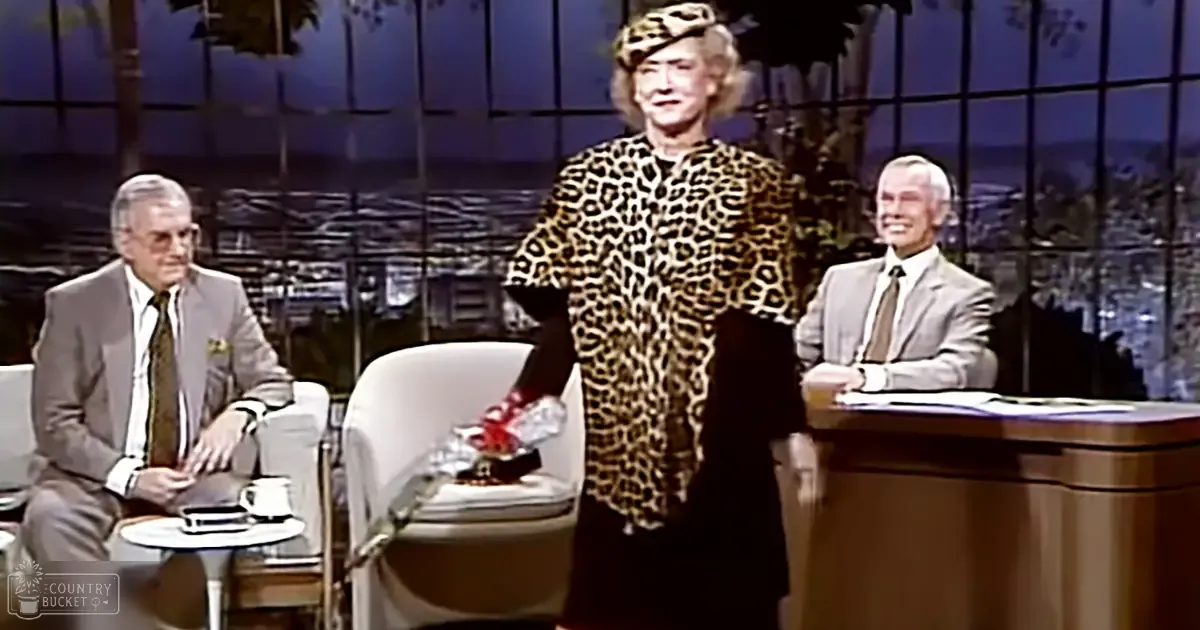An older adult female in her 60s-70s wearing a leopard print jacket and dark dress stands with two other adults, one male middle-aged or older in a light gray suit, on what appears to be a mid-20th century talk show set with urban views through large windows at dusk.