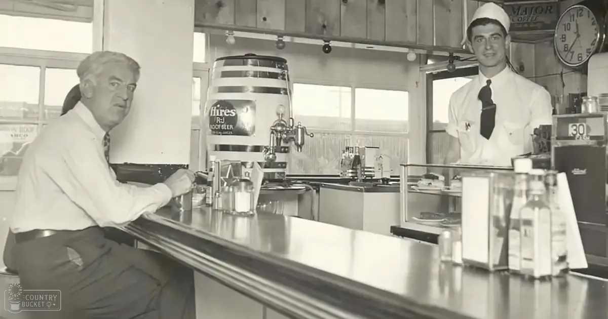 A middle-aged male in a mid-20th century light shirt and tie sits at an old-fashioned diner bar, with a young adult server nearby; the beer tap labeled 'Fires RJ Root Beer' is prominent.