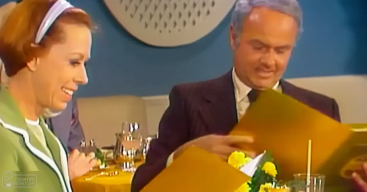 A middle-aged man in a dark suit sits at an indoor table, interacting with a young adult woman wearing a green dress, surrounded by modern decor and artificial lighting.