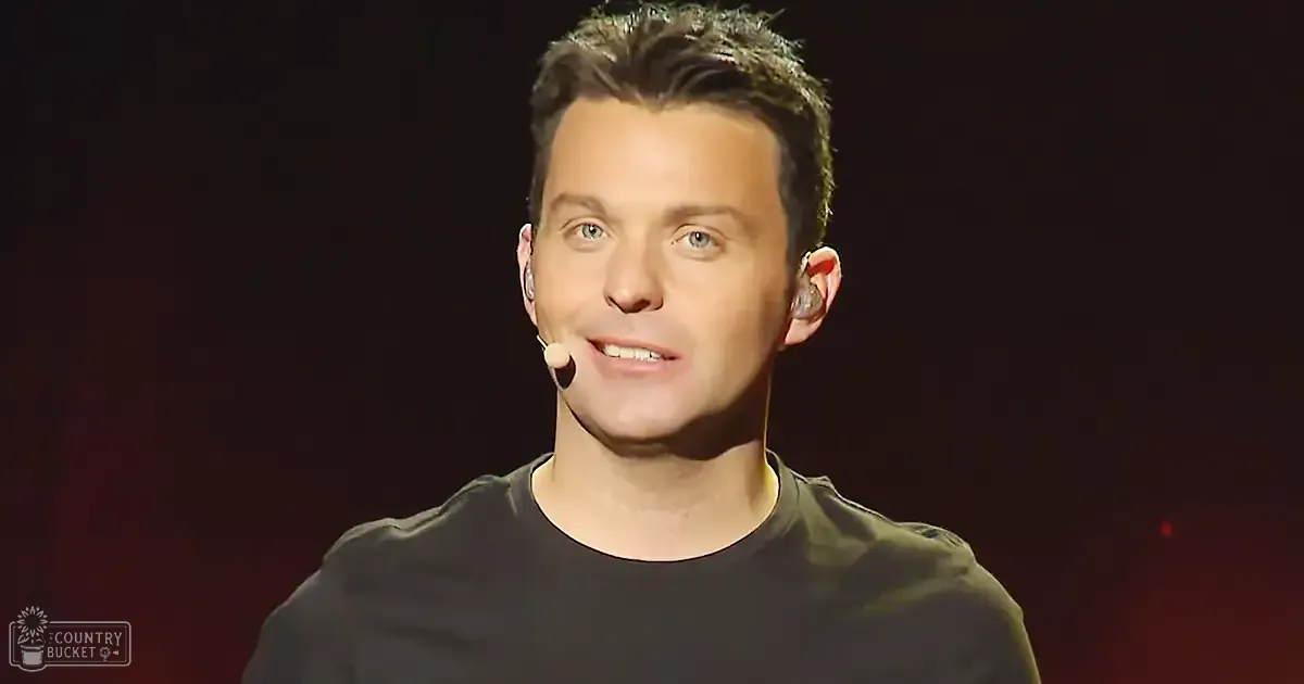 A young adult male in a relaxed fit, dark olive green t-shirt with earbuds and a microphone clipped near his ear, performing on an indoor stage with dark red or maroon tones in the background.