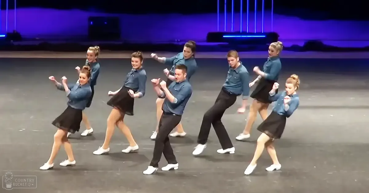 A dance group performing on stage with young adult to adult dancers, male wearing dark blue button-up shirts and black pants, female in knee-length skirts, under indoor venue lighting setup.
