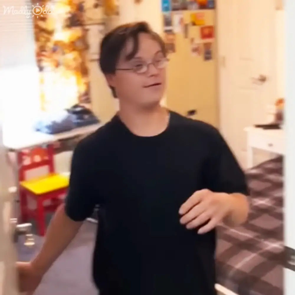 A young adult male wearing glasses stands in a home kitchen or dining area with a yellow chair and tiled floor, surrounded by furniture and decor.