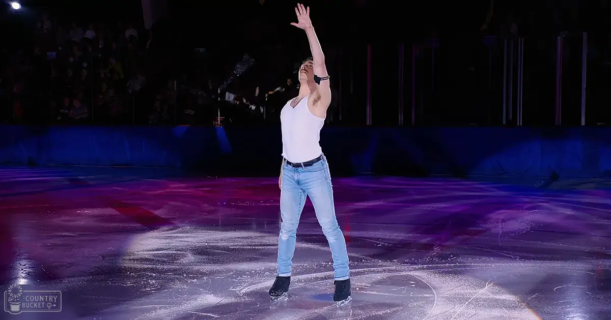 A young adult male figure skating on an indoor ice rink at night with artificial lighting.