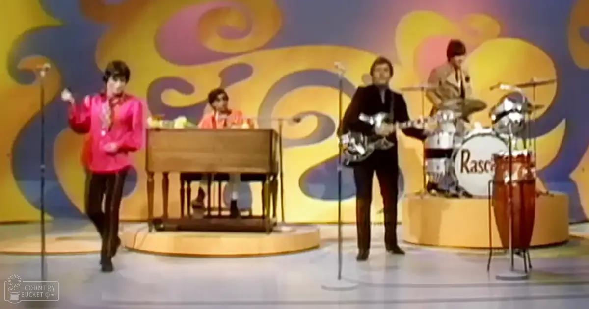 A vibrant, swirling pattern with yellow, orange, and blue colors set against a backdrop of an indoor performance space with microphones and a drum set labeled 'Rascals', suggesting a mid-20th-century style.