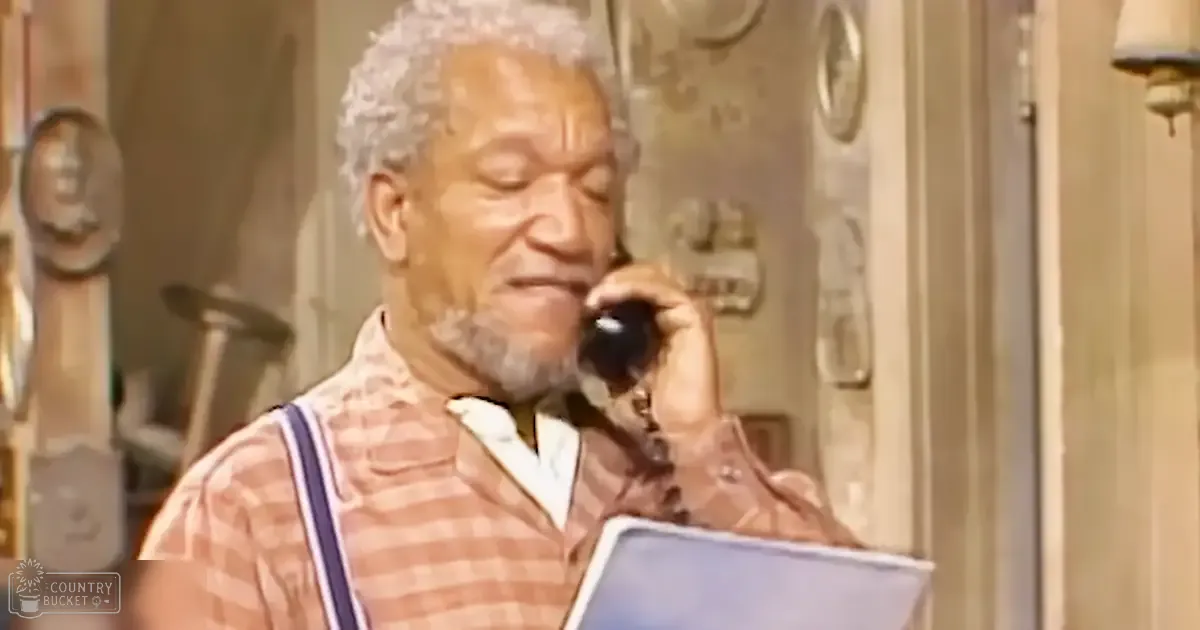 An older adult male, wearing a plaid shirt and suspenders, is on a phone call in a warmly lit home setting with wall decorations.
