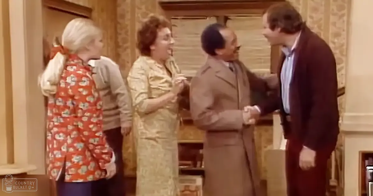 A sitcom scene in a living room with four adults engaged in conversation; the setting includes patterned wallpaper and furniture like a wooden cabinet and fireplace mantel.