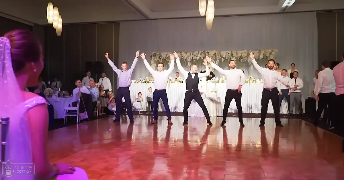 A lively wedding reception with a group of men performing synchronized dance moves, a bride in a white dress partially visible, seated guests in formal attire, and floral decorations indoors under artificial lighting.