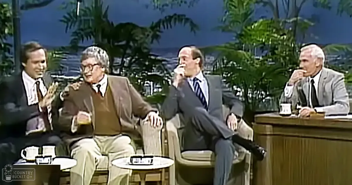 Four male guests seated on a talk show set in an outdoor studio with palm trees and water in the background.