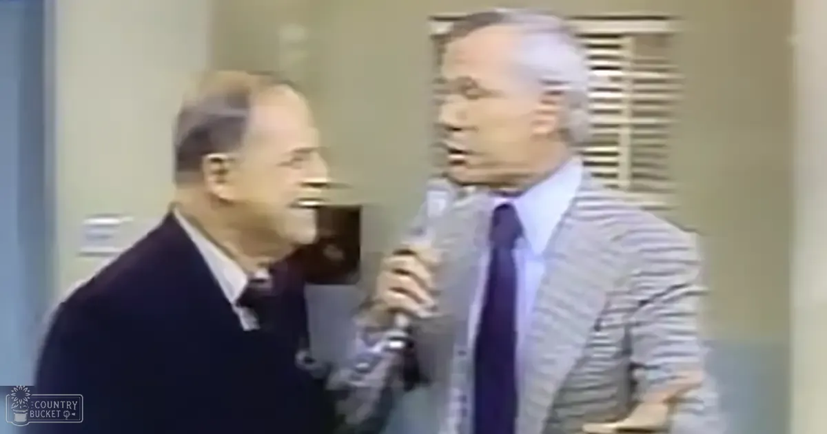 Two middle-aged men in a professional setting, one wearing a dark suit and white shirt with a solid tie, the other in a light gray striped blazer over a light blue shirt and dark purple tie. A handheld microphone is held by Person 2.