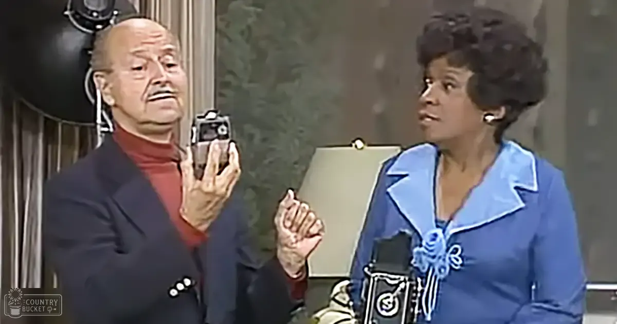 A mid-20th-century living room with an older adult male singer and an adult female actress engaged in conversation, patterned wallpaper and curtains in the background.