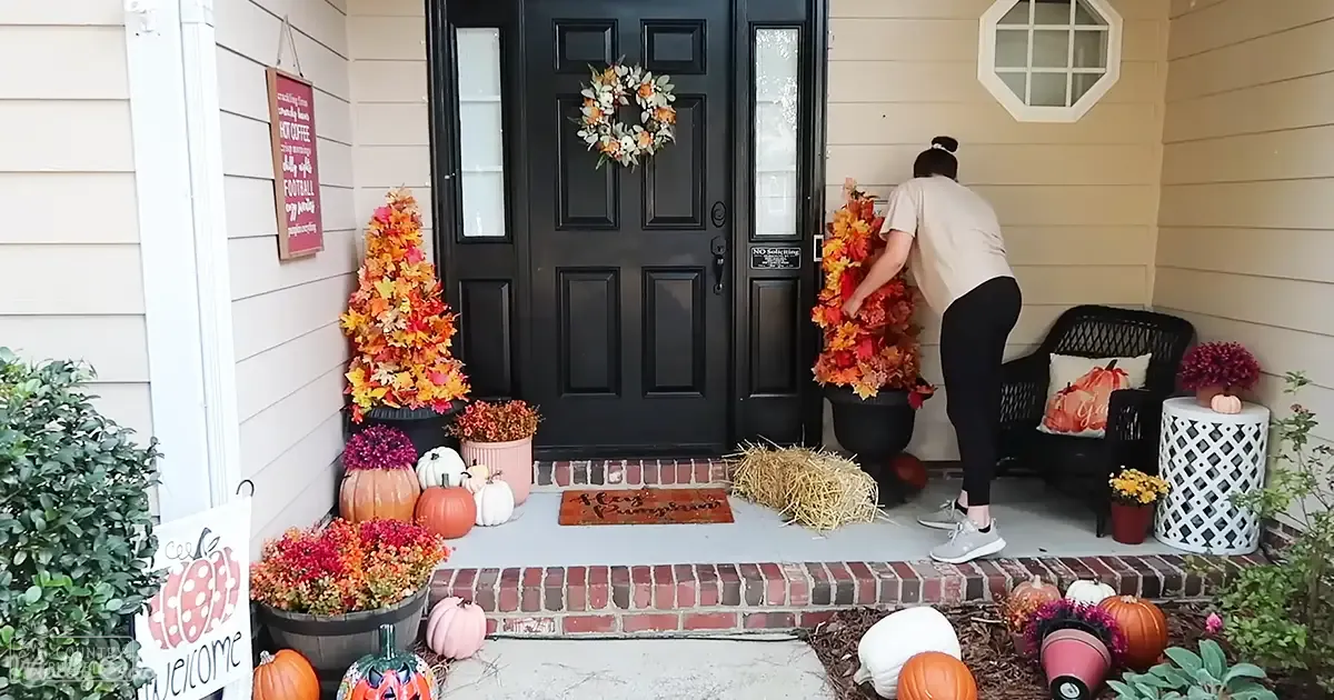 Young adult female decorating a residential porch for fall with autumn-themed decorations, including pumpkins, leaves, hay bales, and fall signs.