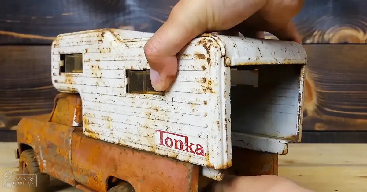 An old, rusted Tonka toy truck with a camper attachment held by a hand against a wooden background.