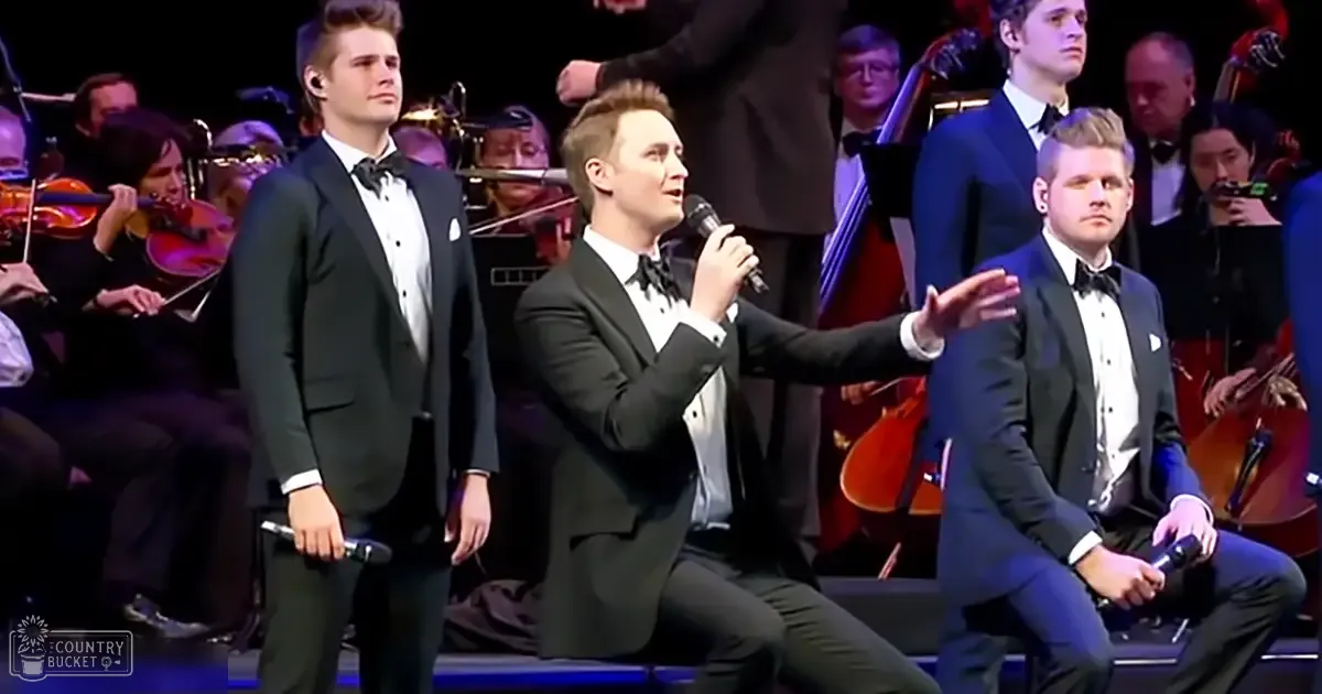 A young adult male singer in a black tuxedo performs on stage with an orchestra, holding a microphone under artificial stage lights.