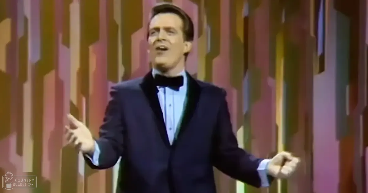 An adult male singer wearing a dark blue tuxedo, white dress shirt, and black bow tie on a stage set with vertical color stripes.