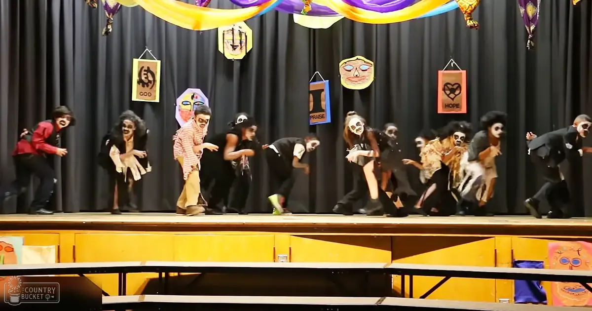 A group of teenagers performing on stage with colorful streamers and signs like 'GOD,' 'HOPE,' and 'PRAISE' in an indoor school auditorium setting, during a performance.