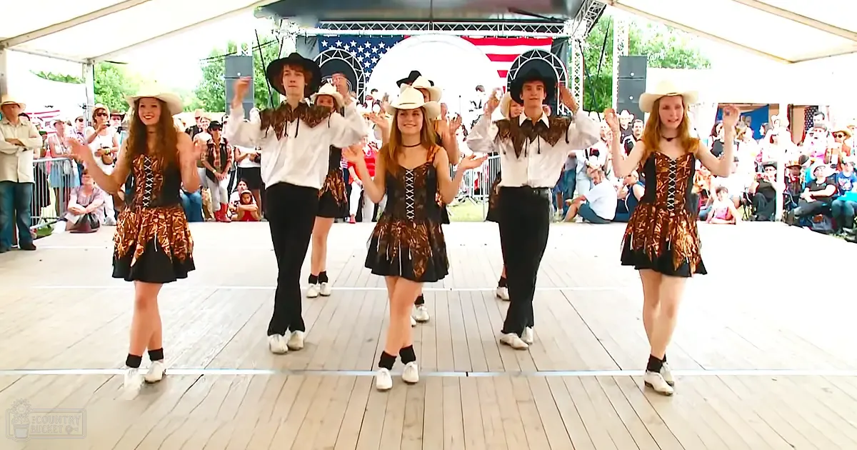 Children and young adults performing Irish dance at an outdoor fairground event with American flag backdrop, spring or summer day, natural sunlight.