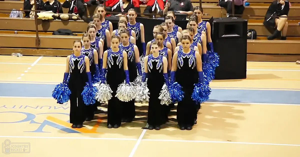 Teenage female cheerleaders in black sleeveless leotards with blue and silver sequins performing at an indoor sports facility, holding blue pom-poms.