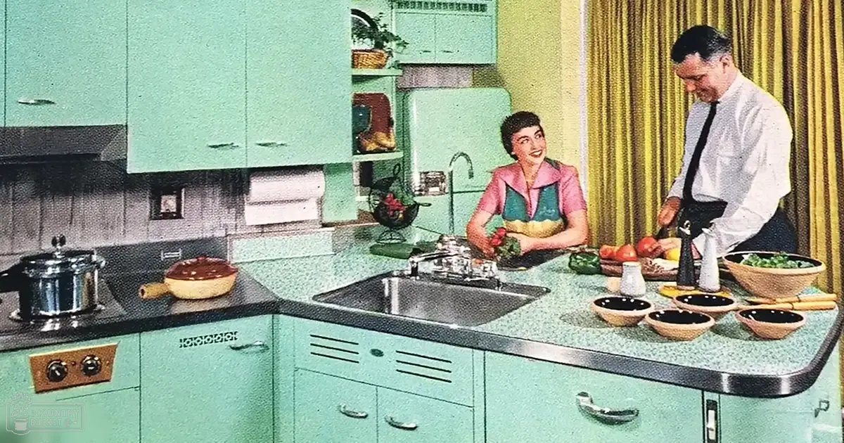 A mid-20th-century kitchen scene indoors, with a woman in a pink and green patterned blouse and a man in a white dress shirt and black tie. The setting includes light teal cabinets with silver handles, a stainless steel sink, and a speckled green formica countertop.