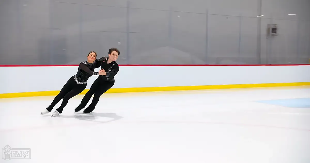 A young adult female figure skater in a black sequined costume and long white figure skates pairs with an adult male figure skater wearing a black fitted jacket and pants, skating on an indoor ice rink with red boundary lines during the daytime.