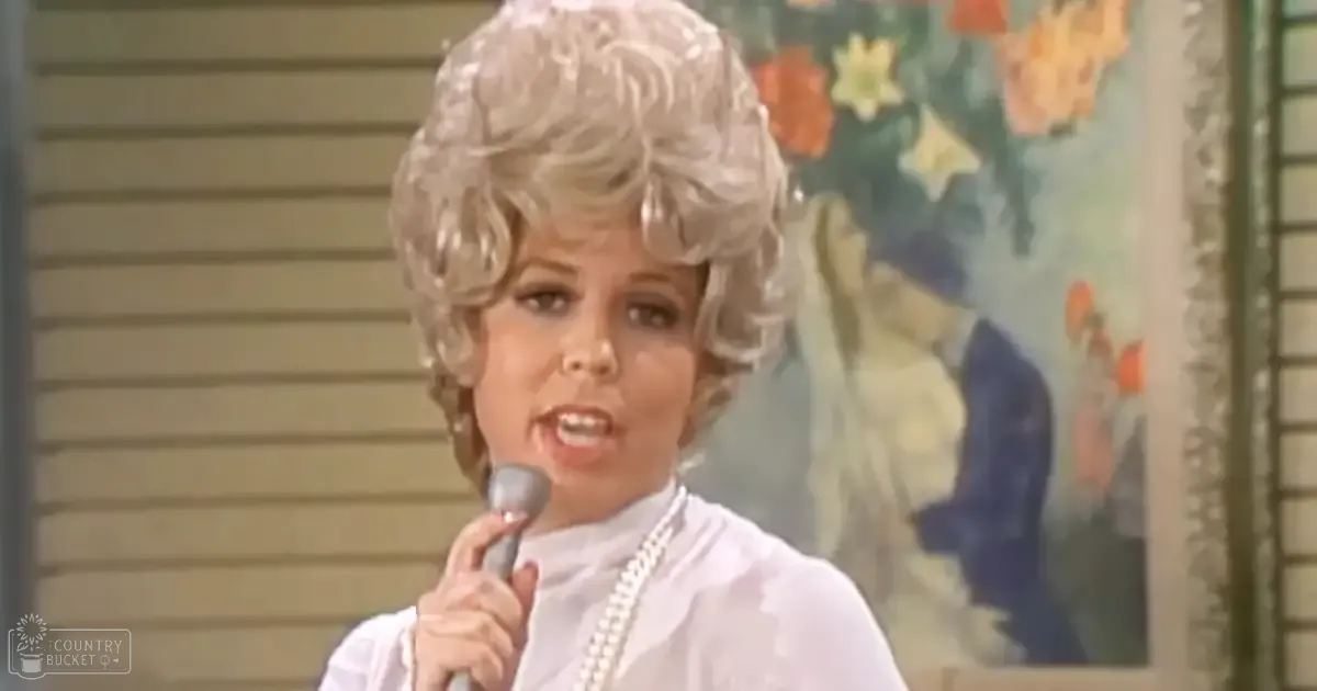 A middle-aged adult female wearing a white, ruffled-collared blouse indoors with natural light and artwork in the background.
