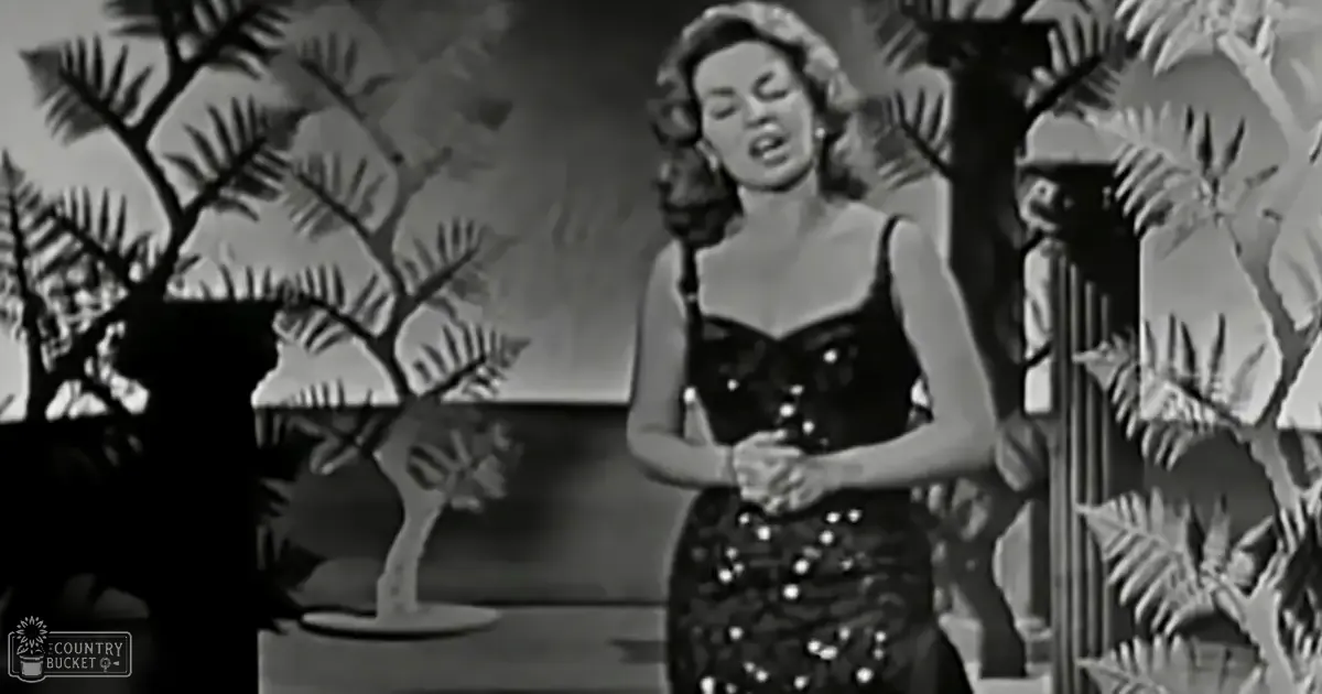 An adult female performer wearing a sleeveless black dress with sequins or beads on stage against stylized tropical foliage in an indoor setting