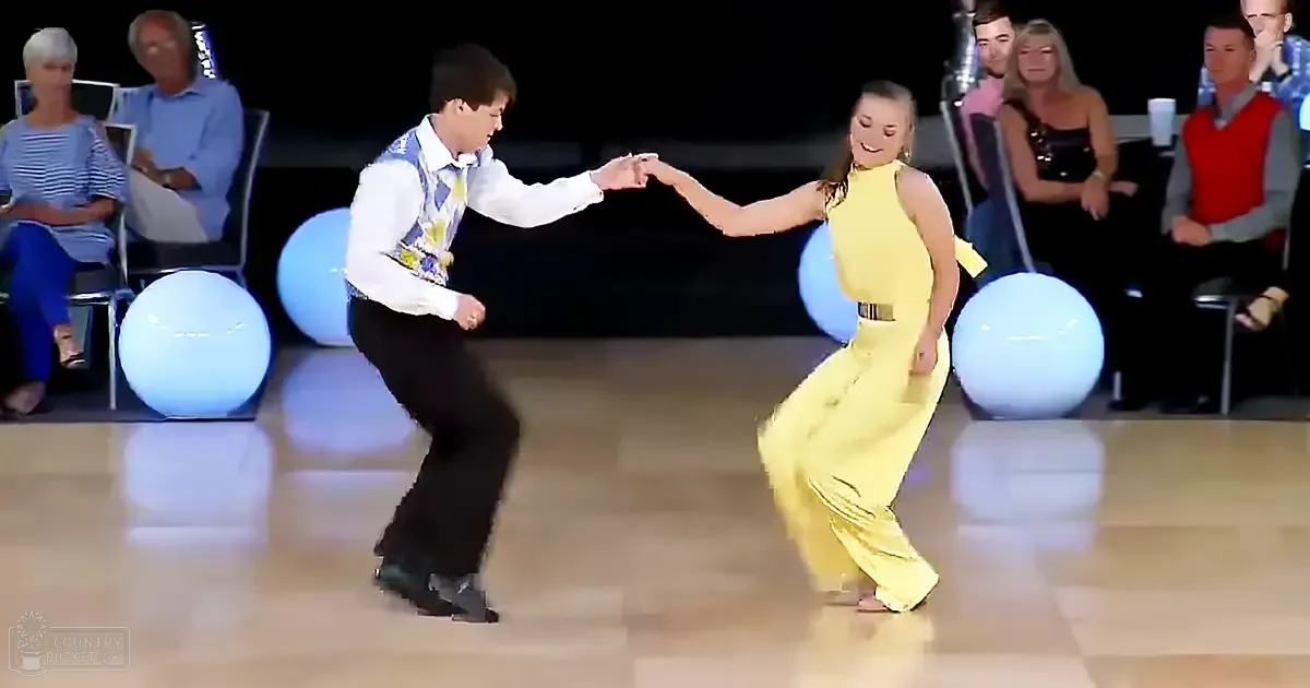 A young adult male and female dance couple performing in a ballroom with seated audience, large spherical light fixtures, and polished wooden dance floor.