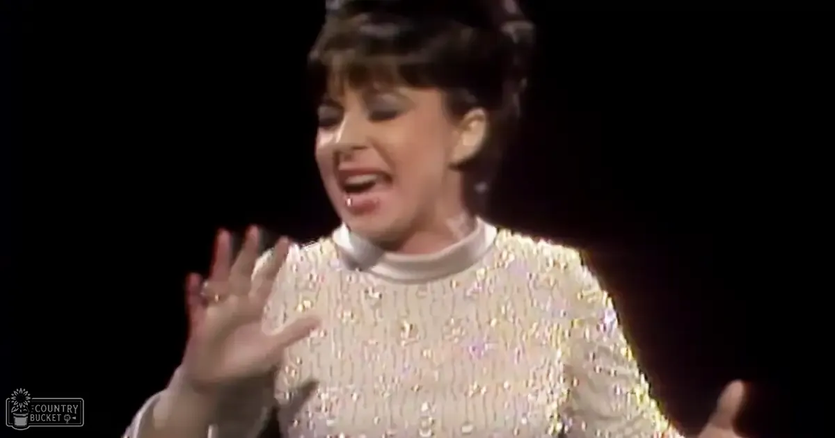 A middle-aged female performer in a white sequined, mid-20th century-style top with long sleeves and a high neckline, wearing a ring on stage under focused lighting.
