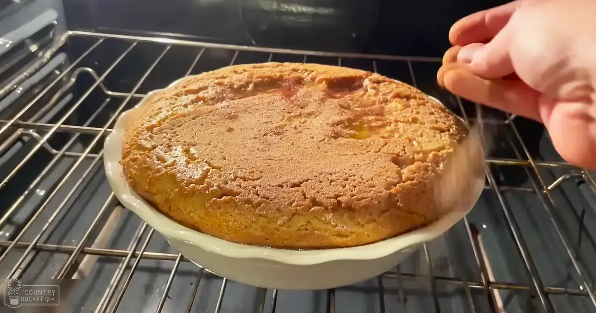 A freshly baked golden brown cake with a slightly cracked surface sits on a cooling rack inside an oven, suggesting it has just been removed from the heat and is cooling down in a home kitchen setting.