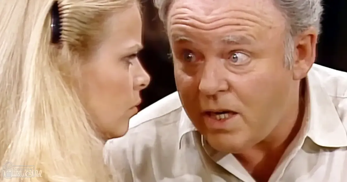 A middle-aged man wearing a light-colored collared shirt stands indoors with a young adult woman whose upper part of her torso is visible, wearing a light-colored blouse or dress; the setting appears to be an indoor environment during evening or night.