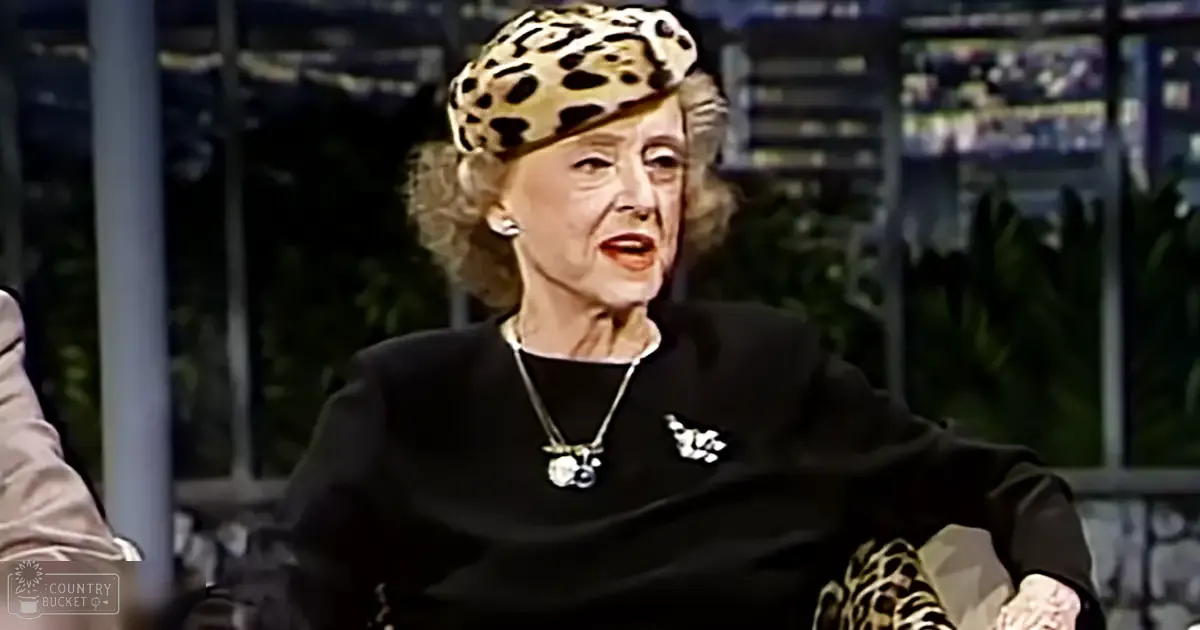 An older adult female in a studio setting, wearing a black dress with long sleeves, a collared neckline, and a leopard print hat; she accessorizes with silver jewelry.