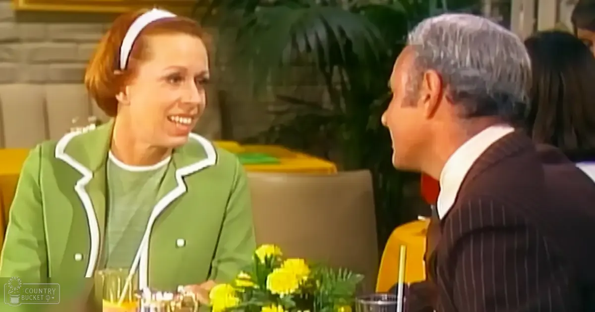A mid-20th-century-style dining scene with an adult female wearing a green jacket and seated at a table adorned with yellow flowers, set against a backdrop of greenery and stone wall paneling.