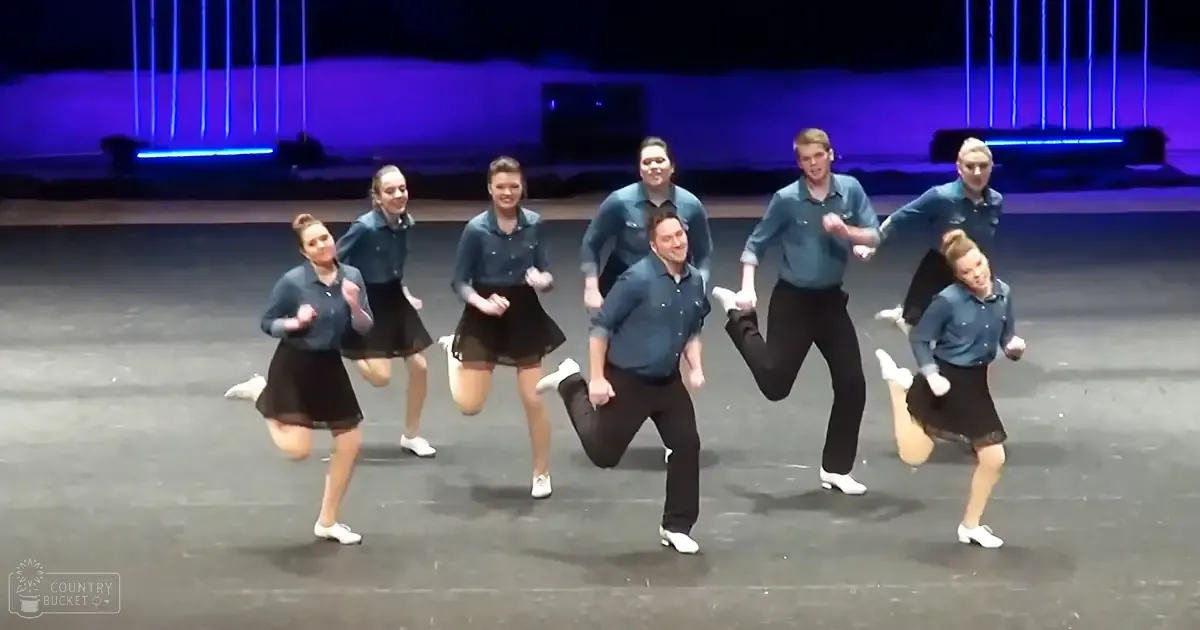 Group performing dance moves on stage with dark blue shirts, black skirts, and white dance shoes under artificial blue lighting.