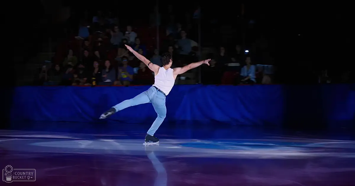 A young adult male figure skater performs gracefully on an ice rink, wearing a white sleeveless tank top, light blue jeans with a belt, and black figure skate boots.