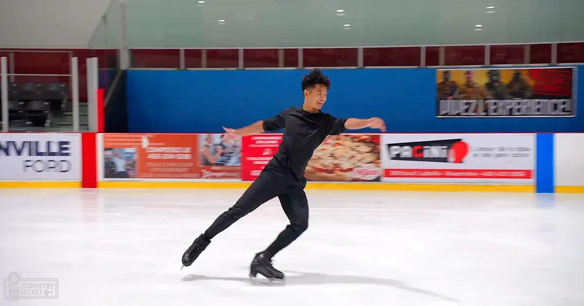 A young adult male figure skating on an indoor ice rink with advertisements on walls, with a black t-shirt and pants.