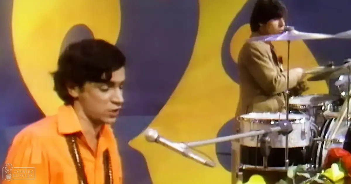 A young adult male singer in an orange shirt performs on stage with a middle-aged male drummer wearing a light brown suit. The backdrop is yellow, suggesting a mid-20th century setting.