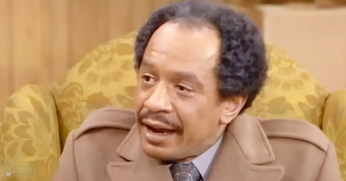 An older adult male seated indoors, wearing a brown blazer and light blue or gray shirt, with yellow floral-patterned upholstery visible behind him.