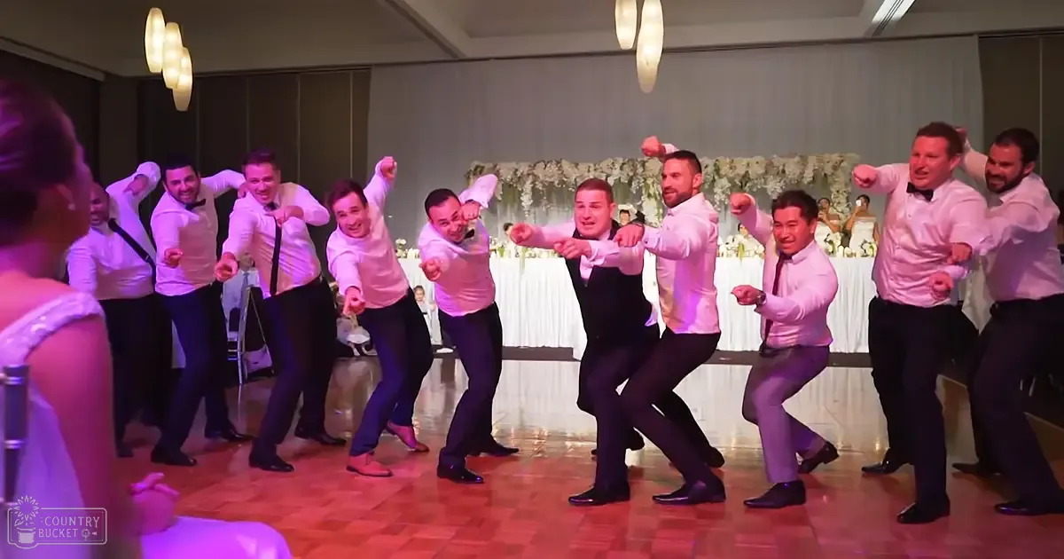 Adult males in formal attire perform a group dance at an indoor wedding reception, with white shirts, black bow ties, dark trousers or tuxedo pants, and beige/cream floral decorations on tables covered in white tablecloths.