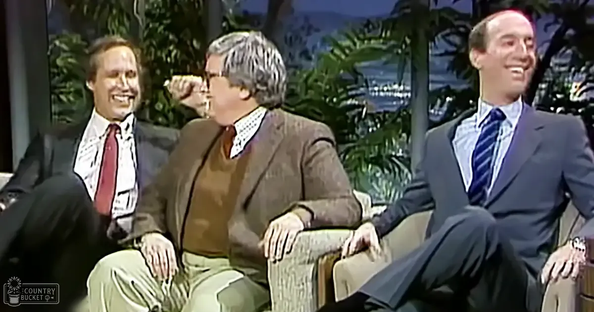 Three men seated on a talk show set in an indoor studio with a backdrop of greenery and water, suggesting an outdoor view used in the background design. The lighting is bright and even, typical of daytime or well-lit indoor conditions.