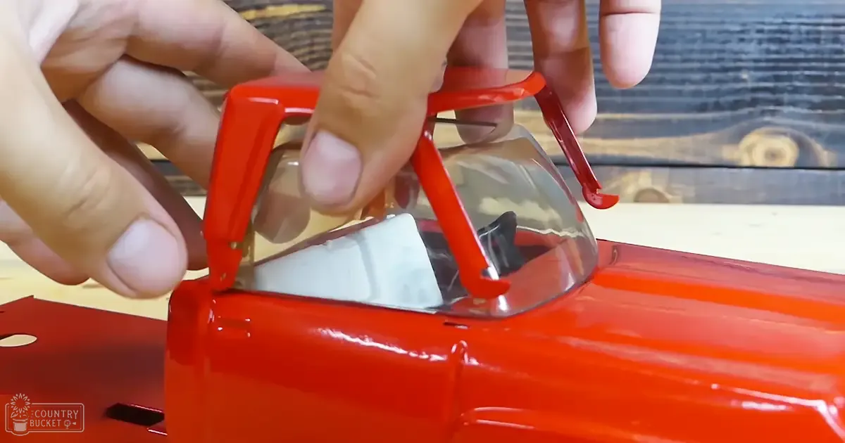 An adult male working on a red toy car with a clear windshield in an indoor setting.