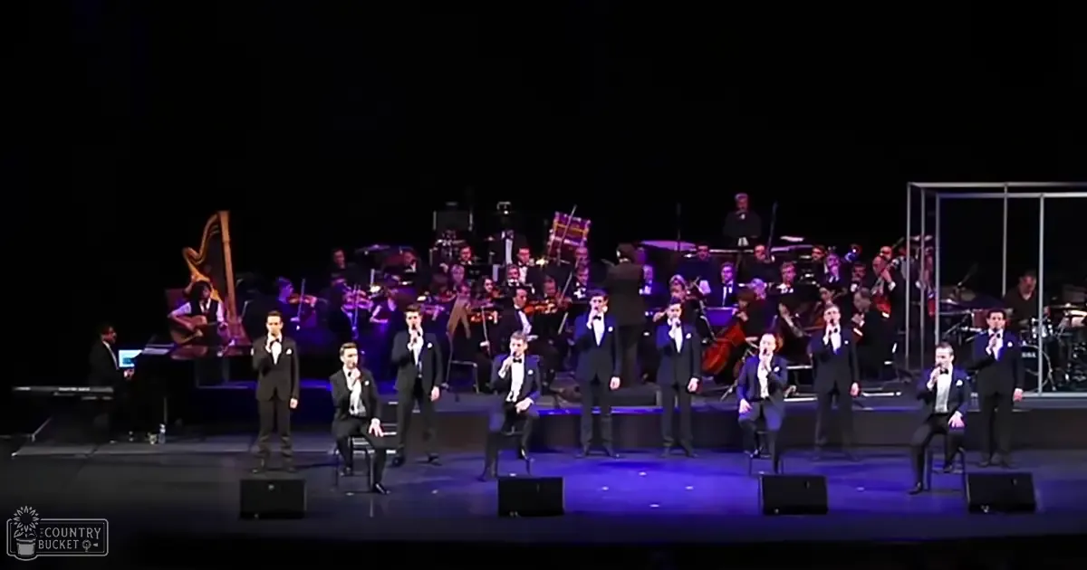 A formal musical performance with young adult male singers in black suits and white shirts, accompanied by an orchestra of adult males dressed similarly, set against the backdrop of a dark curtain on a stage with musical instruments visible.