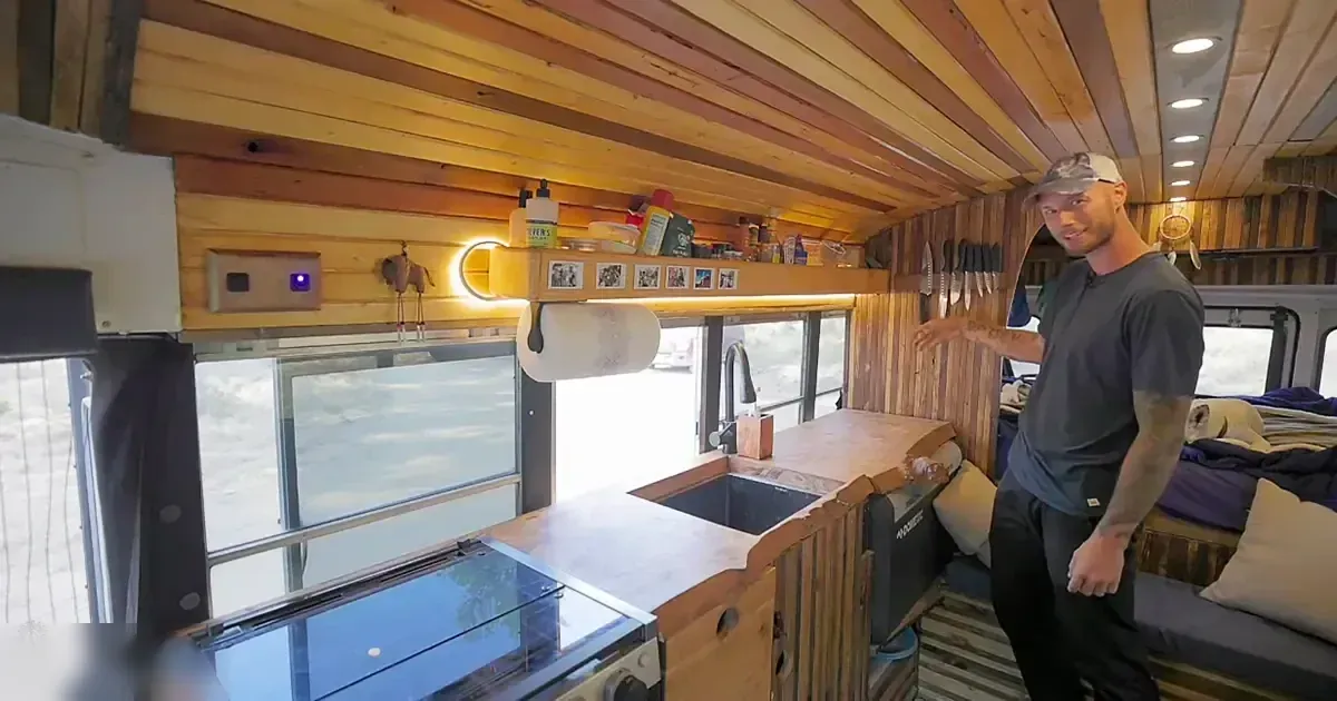 Adult male in a relaxed fit dark gray t-shirt, standing inside a camper van with wooden paneling and natural light streaming through windows.