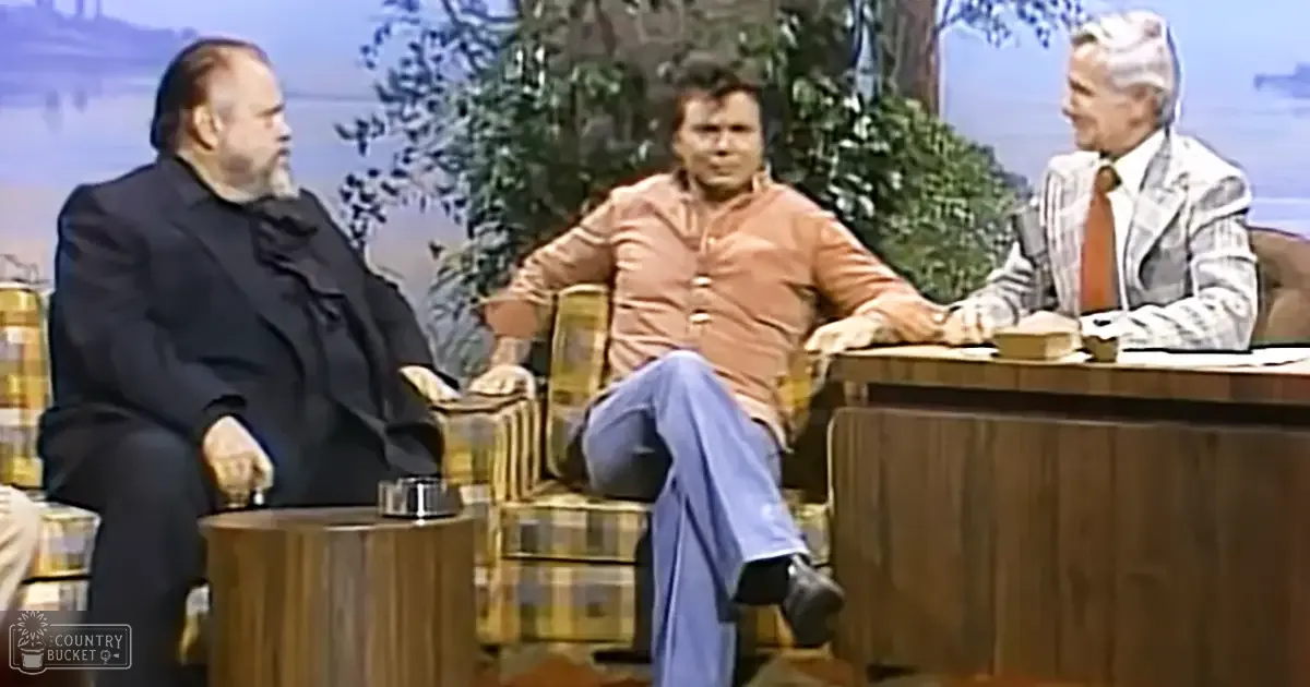 Three adults seated around a wooden table on what appears to be an outdoor set designed for a talk show, with trees and a body of water visible in the background.