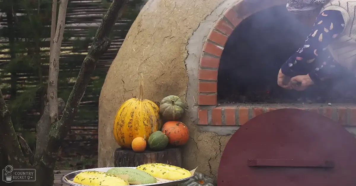 An older adult male wearing a dark-patterned garment and traditional hat, cooking outdoors with pumpkins and gourds displayed near a clay oven in an autumnal setting.