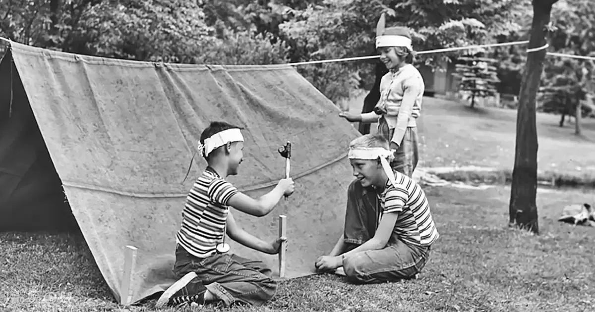 Three children playing near a tent in an outdoor setting with trees and grass in the background; they wear striped shirts, dark pants, and headbands.