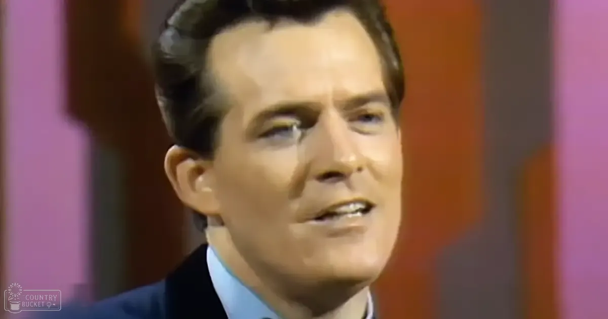 A middle-aged adult male wearing a formal dark suit jacket with a light blue shirt under it stands in an indoor studio setting with colorful panels as the backdrop. The image conveys a professional and polished look, likely for television or film production.
