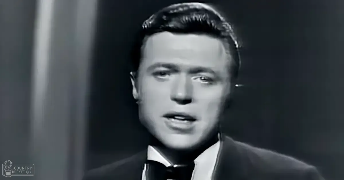 A male adult in a formal suit jacket, shirt with tie, and dark-colored long-sleeved garment stands indoors under controlled lighting.