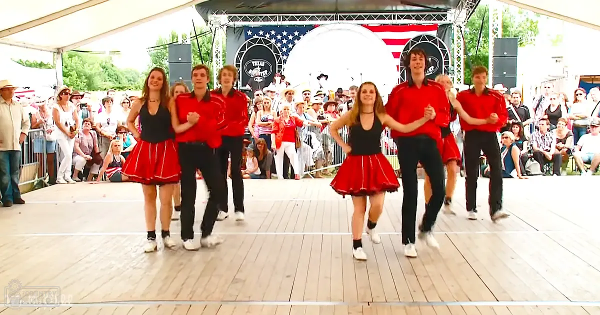 A dance performance at an outdoor event under a large tent, with two young adults wearing matching cheerleading uniforms performing on stage surrounded by an audience dressed casually for spring or summer. The backdrop includes the American flag and 'Texas Frontier' banners.