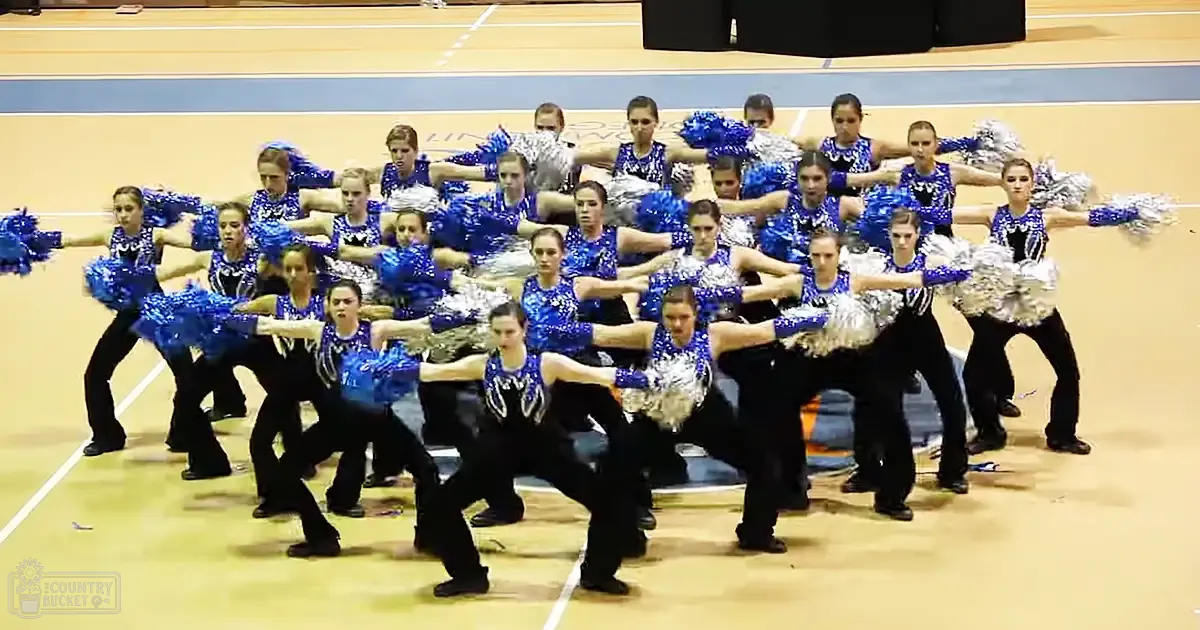 A cheerleading team of young adult females in a well-lit indoor gymnasium performing with blue and black uniforms, metallic silver pom-poms, against a wooden floor backdrop.