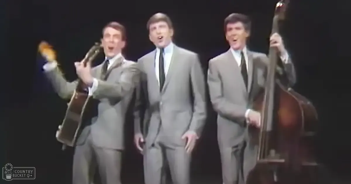 Three adult males in gray suits performing on stage with focused lighting, dark background suggesting an indoor concert hall setting.