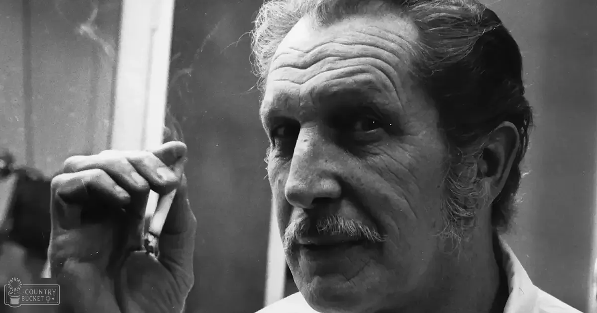 Older adult male in a white shirt outdoors holding a cigarette, with natural light and an urban background.